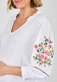 Weiße V-Ausschnitt-Bluse mit Rüschensaum am Ausschnitt und besticktem Blumenmuster am linken Ärmel. Der Stoff wirkt leicht und leicht strukturiert.