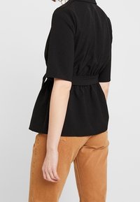 Musta peplum-paita lyhyillä hihoilla, jossa on kerätty vyötärö ja hienovarainen rakenne, yhdistettynä vaaleanruskeisiin housuihin.