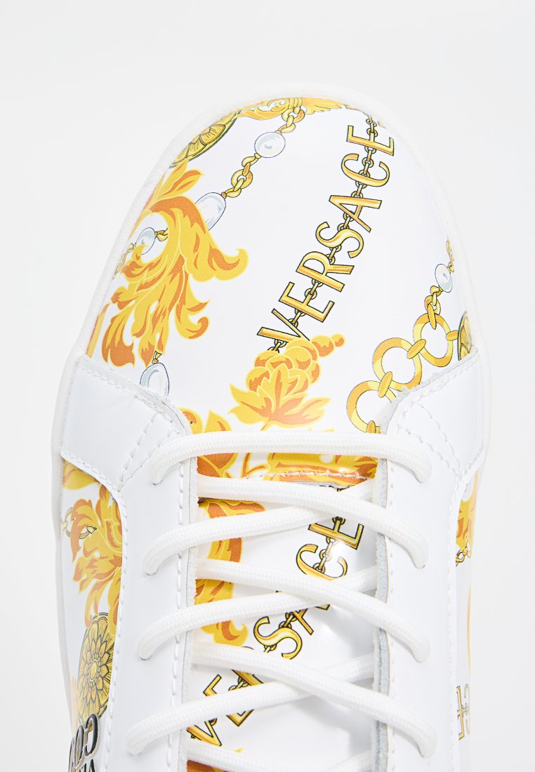 Baskets blanches avec un motif à fleurs jaunes et des éléments de chaîne dorée, accentuées par des lacets blancs.