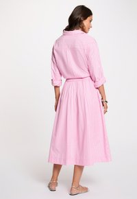 Mujer de espaldas lleva una camisa de manga larga a rayas verticales en rosa y blanco con puños doblados, falda midi a juego, pendientes de aro y sandalias plateadas.