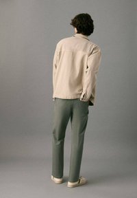 Light beige cotton jacket with a relaxed fit, paired with olive green trousers. Footwear is light-colored with green accents.