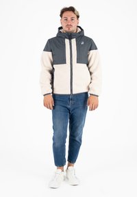 Giacca in pile e nylon, con maniche e parte superiore grigia, corpo crema, zip anteriore, cappuccio e logo sul petto; abbinata a jeans blu e sneakers bianche.