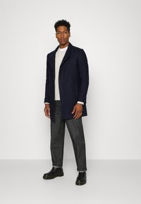 Abrigo navy con un diseño minimalista, cierre de un solo botón, bolsillos laterales, combinado con pantalones a rayas y botines negros.