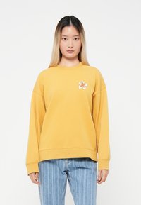 Gul genser laget av bomull, med rund hals og et blomsterdesign på venstre bryst. Kombinert med lyseblå stripete jeans.