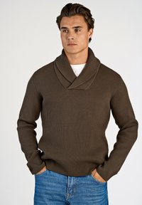 Brauner gerippter Pullover mit Schalkragen, langen Ärmeln und lockerer Passform, getragen über einem weißen Hemd und kombiniert mit blauen Jeans.