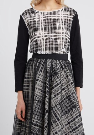 Femme portant un haut noir à manches longues avec un motif en grille abstrait blanc et noir, associé à une jupe large assortie avec un tissu en mesh superposé.
