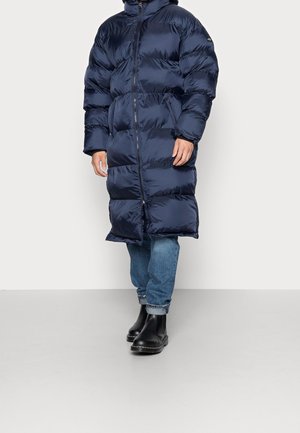 Henkilö, jolla on päällään pitkä tummansininen toppatakki, käännetyt siniset farkut ja mustat nilkkurit, seisoo yksinkertaista vaaleaa taustaa vasten.