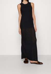 Robe maxi tricotée noire sans manches avec un motif texturé, un décolleté en feston et une fente latérale, associée à des sandales ornées de fourrure.