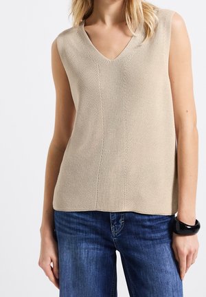 Kvinde iført en beige ærmeløs V-hals striktop med blå jeans og en sort armring på højre håndled, stående foran en hvid baggrund.