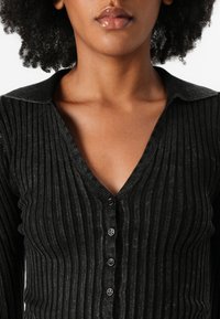 Cárdigan negro de canalé con un profundo escote en V, una tela texturizada y siete botones a lo largo del frente. Diseño con cuello.