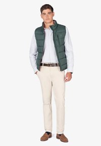 Chaleco puffer verde sobre una camisa clara, pantalones beige y mocasines marrones. El chaleco tiene un cierre frontal y bolsillos laterales. Diseño simple y casual.
