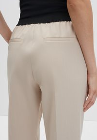 Beige op maat gemaakte broeken met een gladde textuur, voorzien van een gathered elastische tailleband en twee achterzakken. Geen zichtbare patronen of hardware.