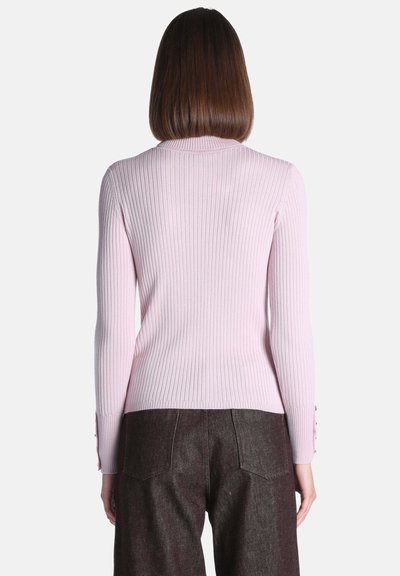 Personne aux cheveux bruns mi-longs portant un pull côtelé rose clair et un pantalon marron foncé, dos au spectateur.