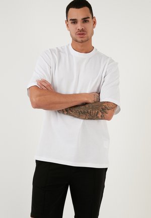 Fehér, rövid ujjú pamut póló, kerek nyakkal. Lazán illeszkedő fazon. A modellen láthatóak a karján a tetoválások, fekete rövidnadrágot visel.