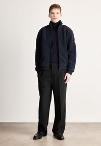 Mereväe fliisjakk kõrge krae ja luku sulgemisega, kombineeritud mustade laiade pükste ja mustade jalanõudega. Sile tekstuur, vabaaja lõige.