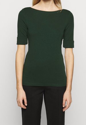 Femme portant un haut moulant vert foncé avec des manches retroussées longueur coude et un pantalon noir, debout devant un fond clair uni.