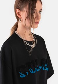 Czarna, oversize koszulka z teksturowanym haftowanym logo "GUCCI" z niebieskimi akcentami płomieni. Model nosi srebrny naszyjnik i kolczyki.