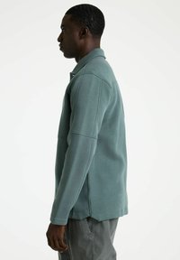 Lichtblauw lange mouw polo sweatshirt met een gestructureerde kraag en zijsplitten, gemaakt van textuurstof, zijaanzicht getoond.