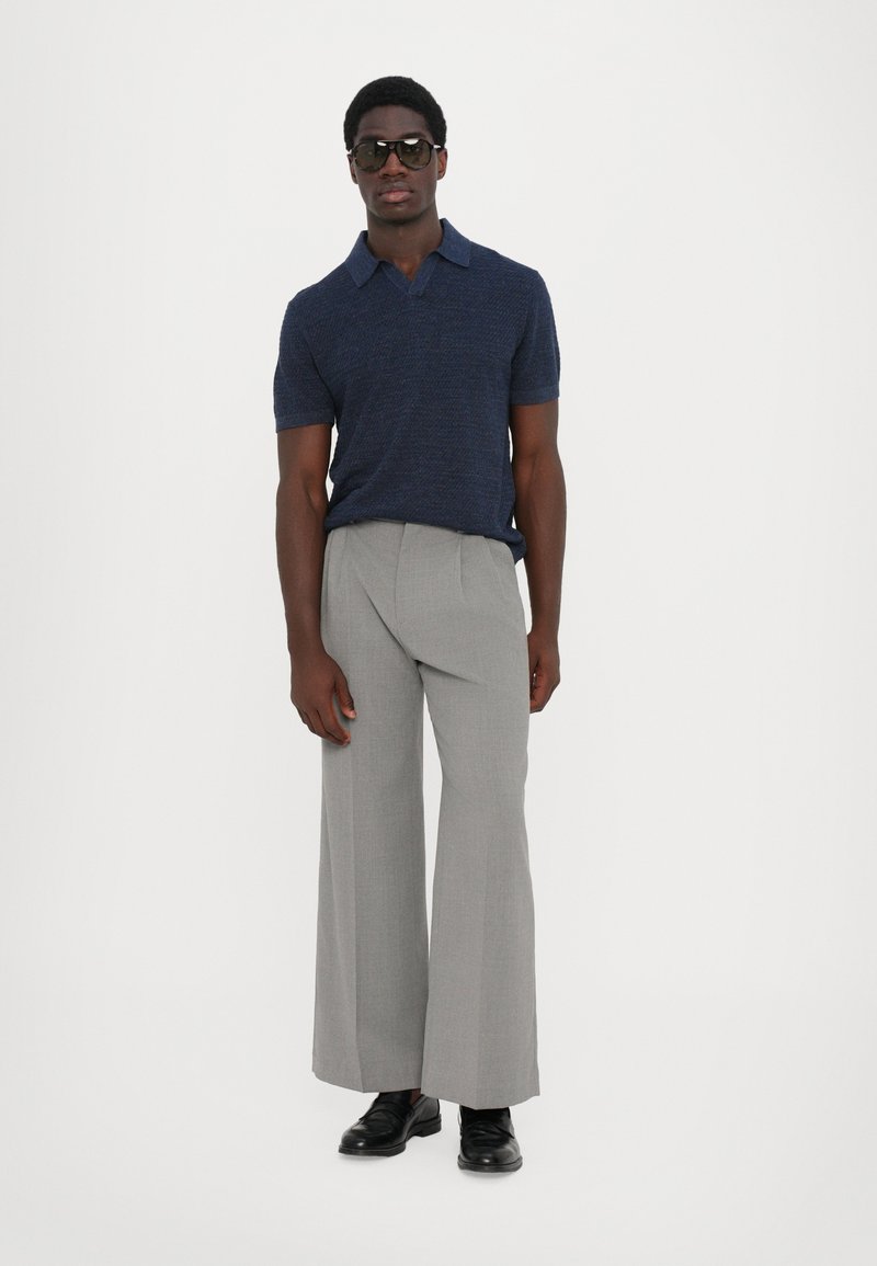 Polo shirt a maglia blu navy, pantaloni a gamba larga grigio chiaro e scarpe nere. Il modello indossa occhiali da sole scuri, con uno sfondo minimale e neutro.