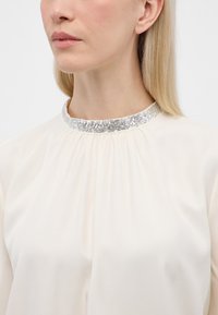 Cremefarvet bluse med et rynket design og en sølvglitrende krave, der viser en blød tekstur og elegant halsudskæringsdetalje.