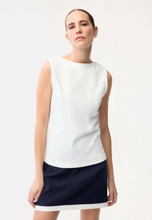 Kobieta w białej bluzce bez rękawów z detalami na szwach oraz granatowej spódnicy z białym obszyciem, stojąca na jednolitym jasnym tle.