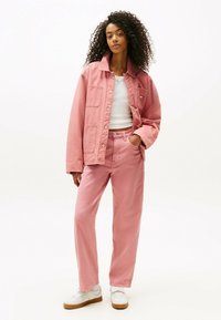 Giacca di jeans rosa chiaro con grandi tasche, abbinata a pantaloni rosa chiaro coordinati. T-shirt bianca sotto e sneaker bianche con suole in gomma.