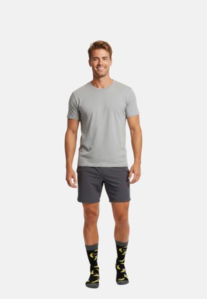 Lächelnder Mann, der ein hellgraues T-Shirt, dunkelgraue Shorts und schwarze Socken mit gelben Bananamustern trägt, vor einem schlichten weißen Hintergrund.