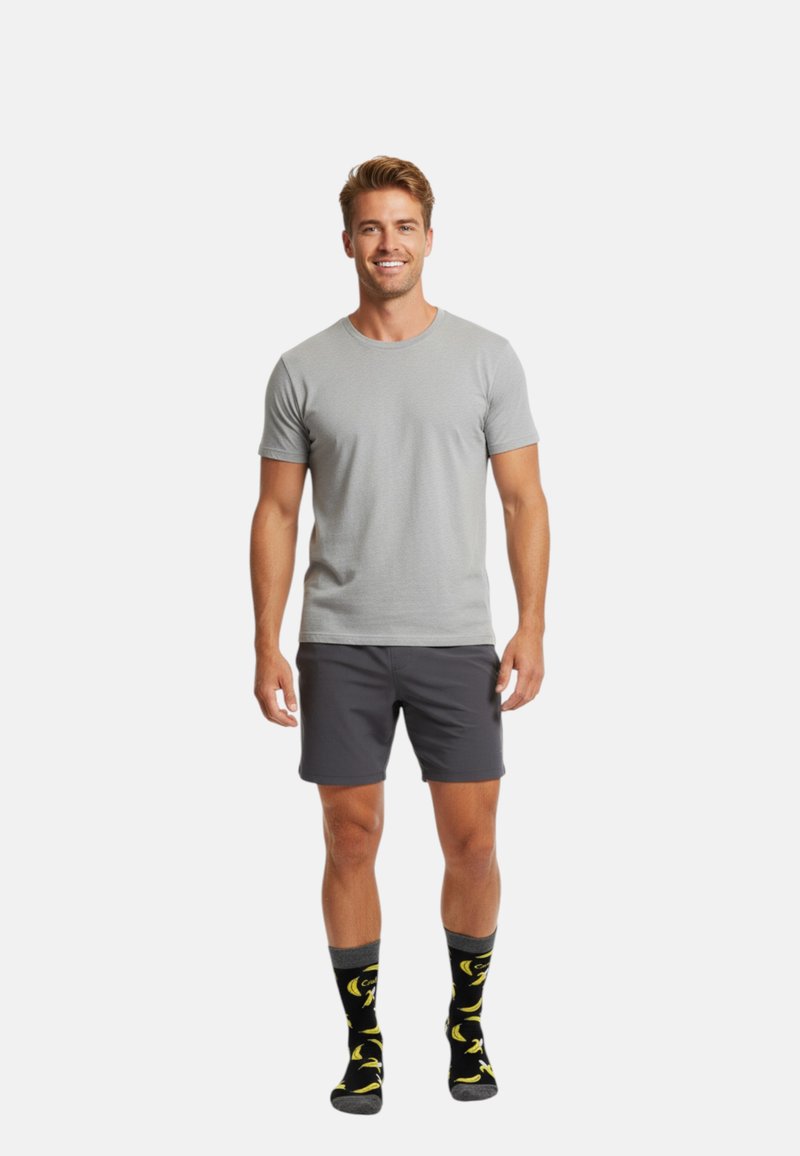 Smilende mand står iført en lysegrå t-shirt, mørkegrå shorts og sorte sokker med gule bananmønstre foran en ensfarvet hvid baggrund.
