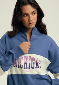 Niebieski i biały sweter z wysokim kołnierzem, zamek błyskawiczny oraz wyraźny tekst "MICHIGAN" w różowym kolorze. Miękka tkanina z prążkowanymi ściągaczami.