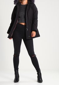 Doudoune noire avec une capuche bordée de fourrure, fermée par des boutons, associée à un crop top gris en tricot côtelé et à un jean skinny noir déchiré, le tout complété par des bottines.