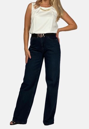 Haut sans manches blanc avec des plis et une accentuation en chaîne dorée, associé à un jean bleu foncé et une ceinture noire avec une boucle en logo doré.