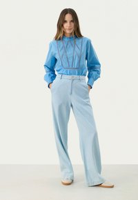 Blauwe katoenen blouse met geborduurde details, gecombineerd met lichte denim wijde broek. Beide kledingstukken hebben een relaxed fit en kraagmanchetten.