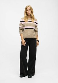 Kootud kampsun beežis, millel on roosa ja mustriibad; omab vabalt langevat lõiget ja rulli mansette. Kombineeritud laia lõikega mustade pükstega ja mustade kingadega.