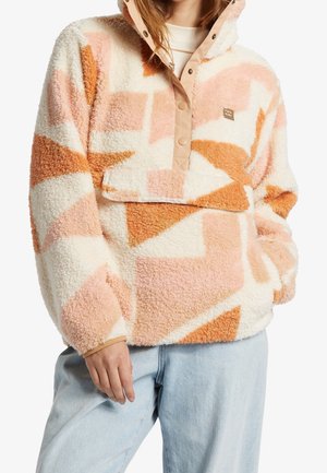 Woman wearing a pastel pink, orange, and white geometric patterned fleece pullover with snap buttons and light blue jeans.