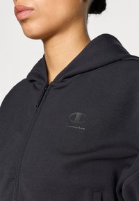 Μαύρο φούτερ με κουκούλα από μαλακό ύφασμα, με φερμουάρ μπροστά, μινιμαλιστικό σχέδιο και τονικό λογότυπο Champion στο στήθος.