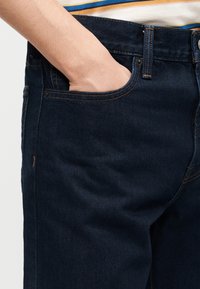 Χέρι που έχει μπει στην μπροστινή τσέπη σκούρου μπλε τζιν, φορεμένο με ριγέ πουκάμισο.