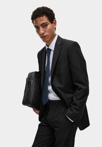 Completo nero con una vestibilità su misura, camicia bianca e cravatta blu, mentre tiene una borsa in pelle nera. Texture liscia e bottoni eleganti, sfondo grigio neutro.