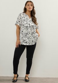Donna in piedi contro un muro semplice, che indossa una blusa bianca con stampa floreale nera, pantaloni skinny neri e scarpe basse nere.