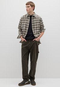 Kariertes beige-schwarzes Flanellhemd über einem schwarzen T-Shirt, kombiniert mit olivgrünen Hosen und dunklen Turnschuhen.