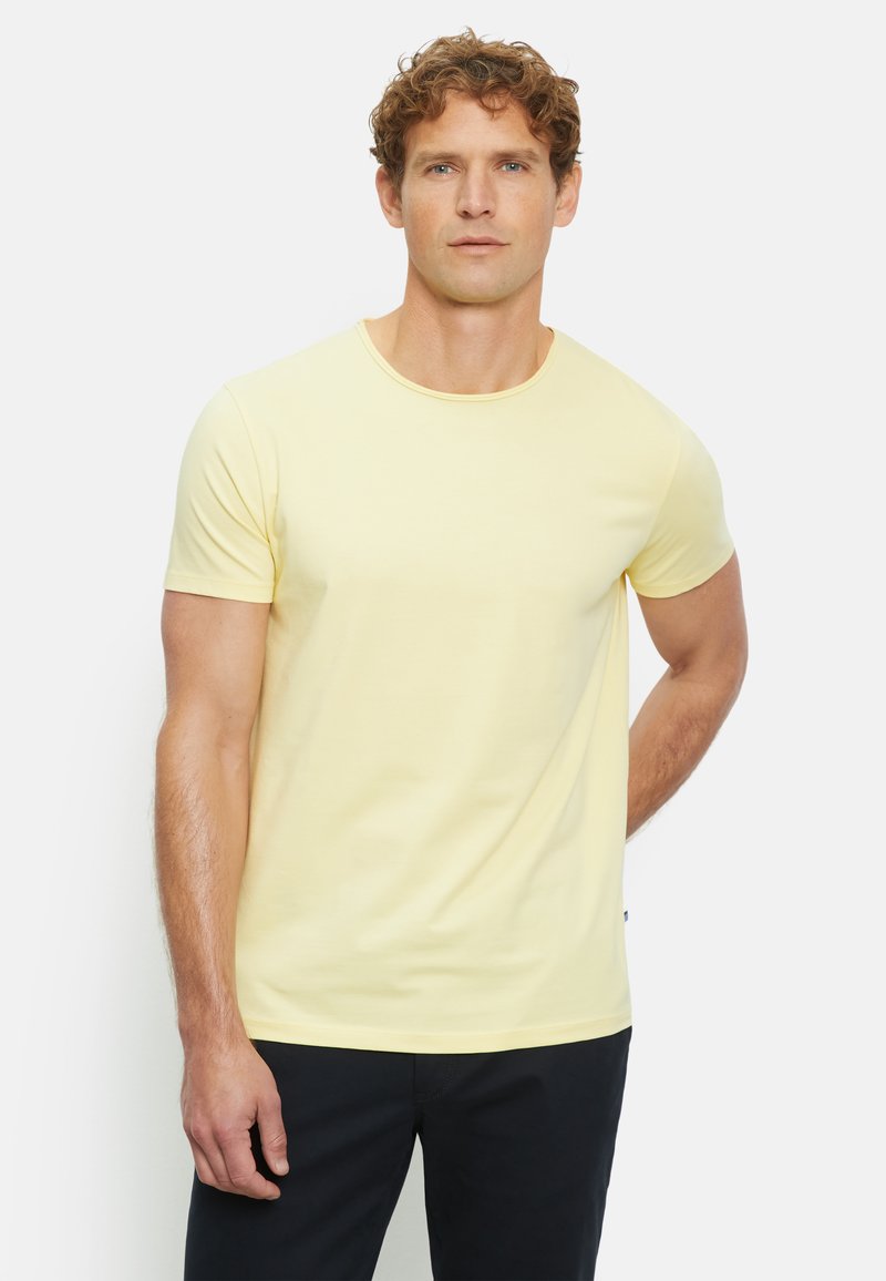 T-shirt gialla in cotone con colletto rotondo, maniche corte e una texture liscia, abbinata a pantaloni scuri, su uno sfondo semplice.