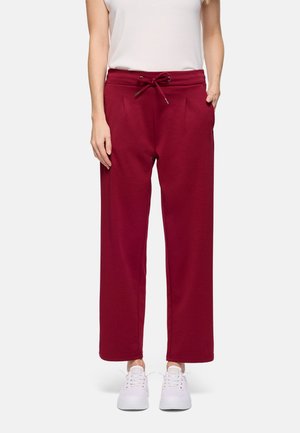 Mujer con pantalones deportivos rectos color burdeos con cintura ajustable mediante cordón, bolsillos laterales, zapatillas blancas y camiseta blanca sin mangas.