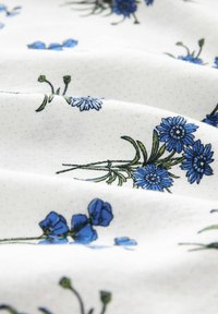Tyg med blå blommig broderi på vit bakgrund. Mönstret inkluderar olika blommor och blad med en mjuk textur.