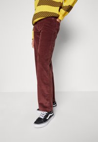 Corduroy trousers in dark red, featuring a ribbed texture and a straight-leg design. Paired with black sneakers and a yellow striped sweater.