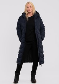 Cappotto imbottito blu navy, design lungo con cappuccio, texture trapuntata, chiusura frontale con zip e tasche laterali, indossato sopra una maglietta nera e pantaloni.