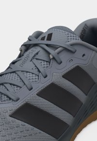 Κοντινή λήψη γκρι αθλητικού παπουτσιού Adidas με μαύρες τρεις ρίγες, πλεκτό επάνω μέρος και το κείμενο «DROPSET» κοντά στα κορδόνια σε λευκό φόντο.