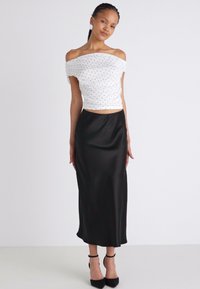 Bela off-shoulder majica s črnimi pikami, v kombinaciji s črno satensko midi krilom in črnimi petami s trakovi okoli gležnja.