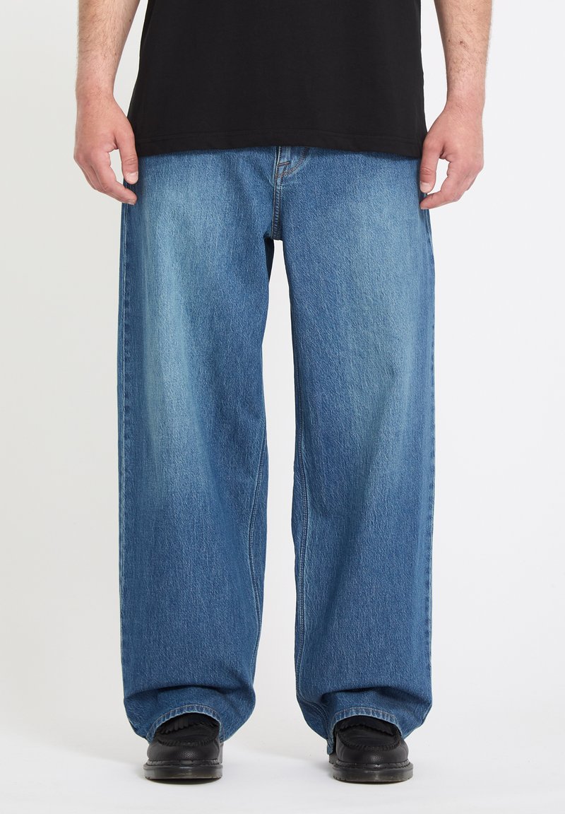 Homme portant un jean bleu ample, une chemise noire et des chaussures noires, debout devant un fond blanc uni.