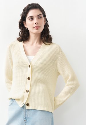 Frau mit dunklem lockigem Haar, die eine cremefarbene Strickjacke mit Knöpfen über einem weißen Oberteil und hellblauen Jeans trägt, blickt leicht nach rechts.