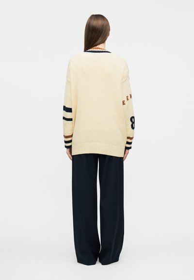 Άτομο με μακριά καστανά μαλλιά στέκεται γυρισμένο προς τα πίσω, φοράει πουλόβερ σε κρεμ χρώμα με ρίγες και αριθμούς στα μανίκια και μαύρο παντελόνι με φαρδύ πόδι.