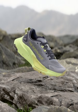 Grijze en neon groene The North Face trailschoen zwevend boven rotsachtig terrein met vervaagde bergen op de achtergrond.